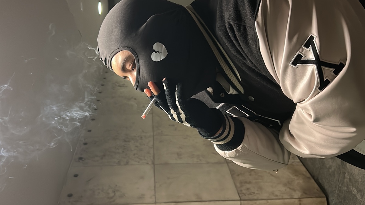 Big Z in a black broken-heart balaclava and varsity jacket, smoking against a tiled wall